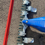 Close-up of a blue agricultural implement with toothed metal prongs mounted on a black frame, lying on rough asphalt beside a red beam piece.