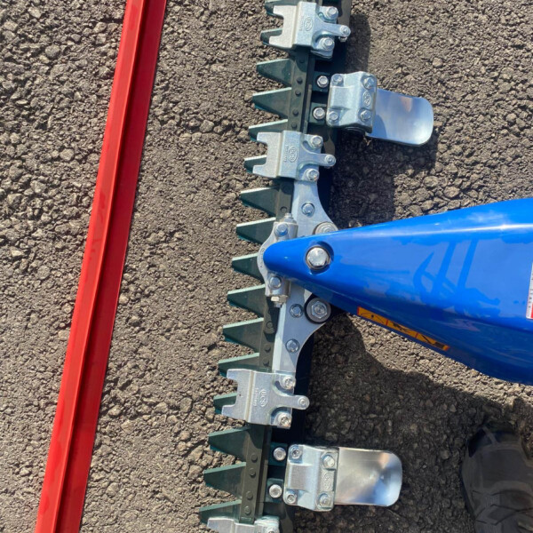 Close-up of a blue agricultural implement with toothed metal prongs mounted on a black frame, lying on rough asphalt beside a red beam piece.