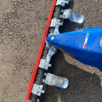 Close-up of a blue construction machine with a red cutting edge blade and silver mounting brackets on rough ground.