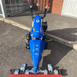 Blue BCS garden tractor with a wide red sweeper attachment parked on a driveway near a brick house and metal gate.