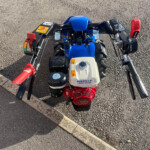 From a rider's viewpoint: blue mini ATV handlebars with red grips and a visible engine, sitting on asphalt beside a curb.