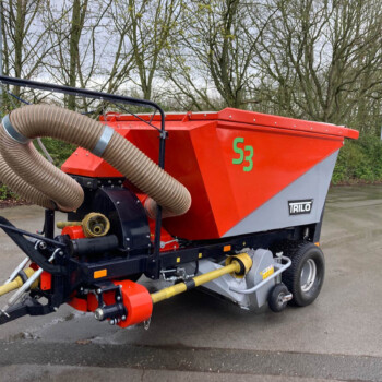 Red agricultural spreader with large corrugated hose attached, branded TRILO, parked on a paved area with trees in the background