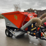 Orange mobile concrete mixer trailer with a long beige hose connection, parked on a wet paved lot.