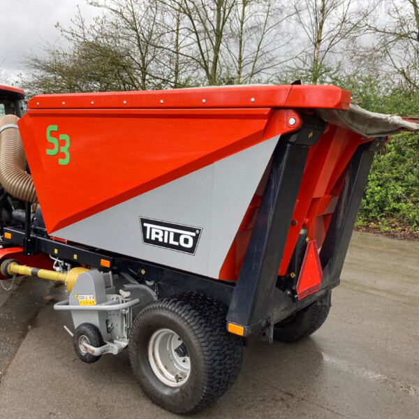Red TRILO manure/seed spreader attached to a tractor, parked on a wet paved surface with trees in the background.