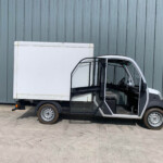 White delivery van with its front passenger door open beside a teal-gray corrugated metal wall.