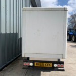 Rear view of a white box trailer attached to a trailer, with blue tractors visible in the distance beside a corrugated metal building under a blue sky.