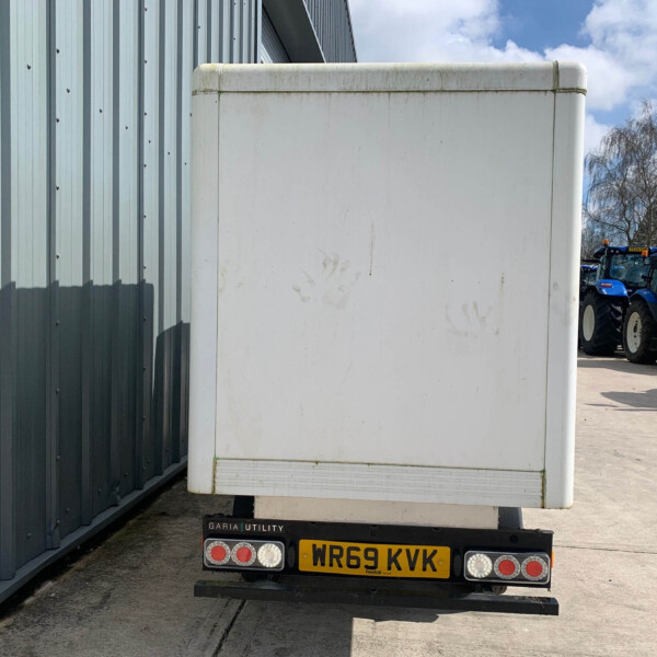 Rear view of a white box trailer attached to a trailer, with blue tractors visible in the distance beside a corrugated metal building under a blue sky.