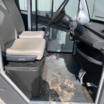 Interior of a cabin with beige seats and a black steering wheel; keys in ignition, dusty floor with leaves visible outside the cab door.