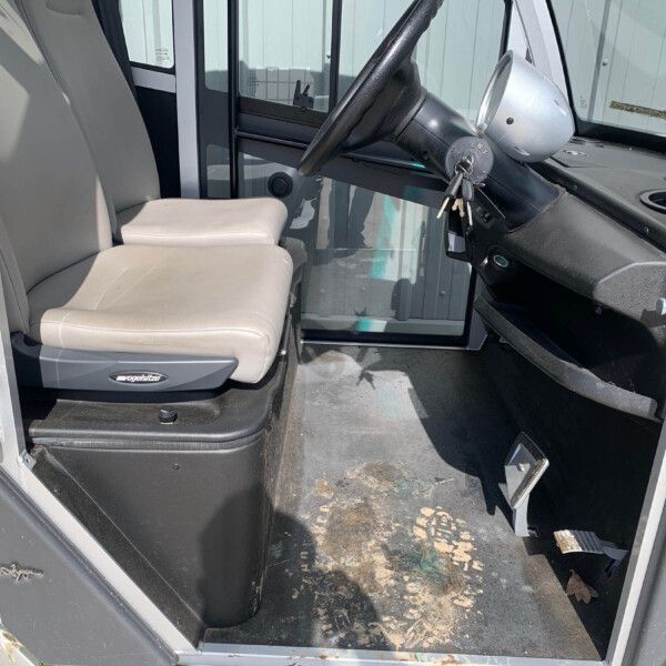 Interior of a cabin with beige seats and a black steering wheel; keys in ignition, dusty floor with leaves visible outside the cab door.