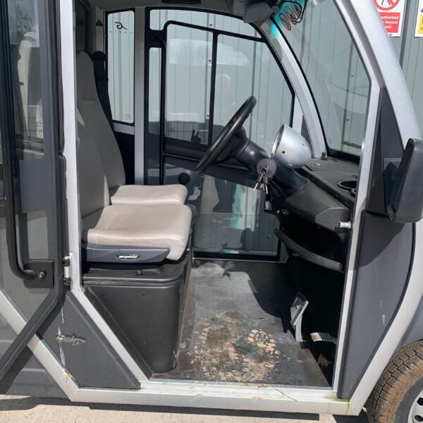 Interior view of a truck cab with a beige driver’s seat, steering wheel, and open door; exterior signs Behind the cab are visible on a metal wall.