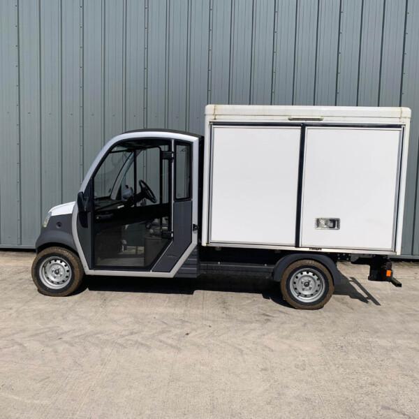 White delivery van with the driver’s door open, parked in front of a tall gray corrugated-metal wall.