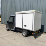 White box truck parked beside a gray corrugated metal warehouse wall, with a security camera and warning signs nearby.