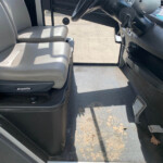 Interior of a bus cab: two light gray driver seats, steering wheel and dashboard on the right, keys hanging, worn floor with leaves and dirt