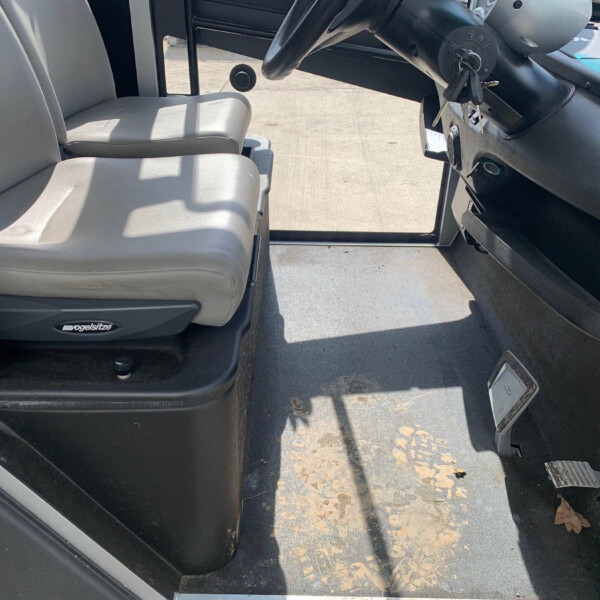 Interior of a bus cab: two light gray driver seats, steering wheel and dashboard on the right, keys hanging, worn floor with leaves and dirt