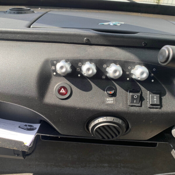 Center console of a car with climate knobs, hazard button, and a small 'Body Light' indicator; a few papers lie in the lower tray.