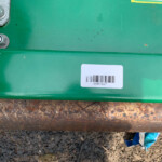 Close-up of a green metal surface with two large bolts on the left and a white barcode label, showing dirt and rust along the bottom edge.