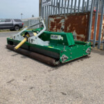 Green street-sweeper machine parked on an asphalt surface beside a rusted metal cabinet and a tall metal fence. In the background, a silver pickup truck sits on the curb.