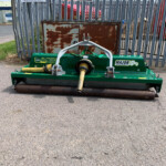 Green Major mower attachment with yellow drive shaft sits in front of a rusty cabinet behind a tall metal fence in an industrial yard.