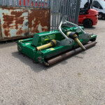 Green road roller attachment in the foreground with a rusted metal cabinet and a chain-linked fence behind it; a red truck and white van sit in the background in a parking lot area.
