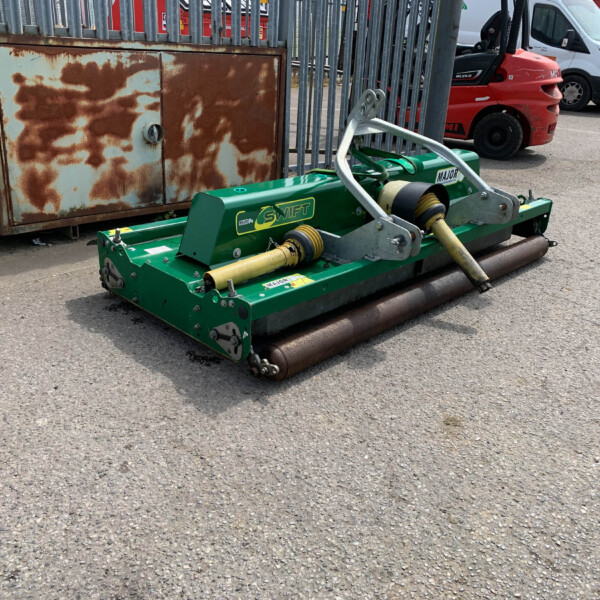 Green road roller attachment in the foreground with a rusted metal cabinet and a chain-linked fence behind it; a red truck and white van sit in the background in a parking lot area.