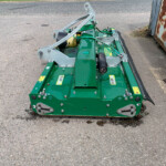 Green agricultural machinery attachment with a metal frame and hydraulic hoses resting on a paved surface; likely a mower or conditioner implement.