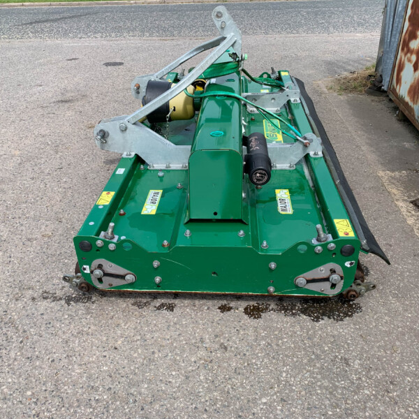 Green agricultural machinery attachment with a metal frame and hydraulic hoses resting on a paved surface; likely a mower or conditioner implement.