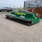 Green industrial street sweeper attachment on asphalt, with a metal fence and a parked SUV in the background.