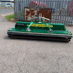 Green street sweeper parked on a paved area in an industrial yard with a rusted metal cabinet and fence in the background.