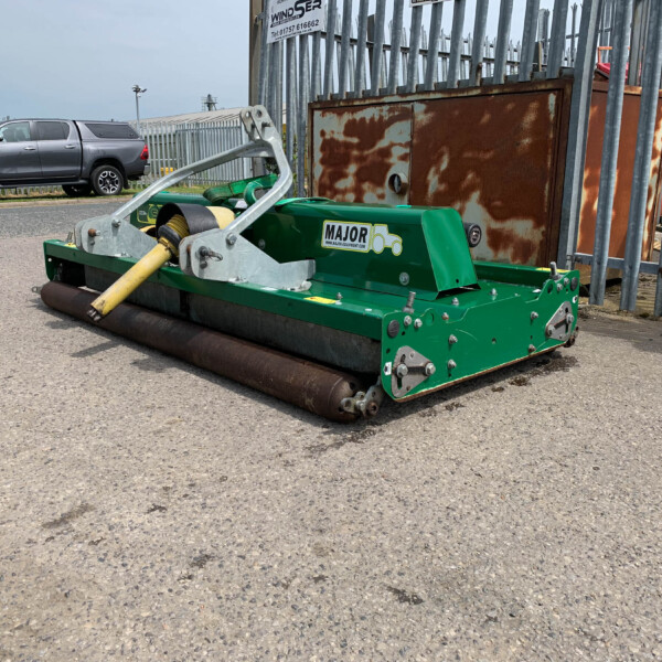Green street-sweeper machine parked on an asphalt surface beside a rusted metal cabinet and a tall metal fence. In the background, a silver pickup truck sits on the curb.