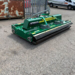 Green street-sweeping machine with a roller brush parked on an asphalt lot, rusted metal cabinet behind.