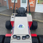 Front view of a small off-road vehicle with a black roll cage, large knobby tires, and a Russell's logo on the hood, parked outside a brick building.