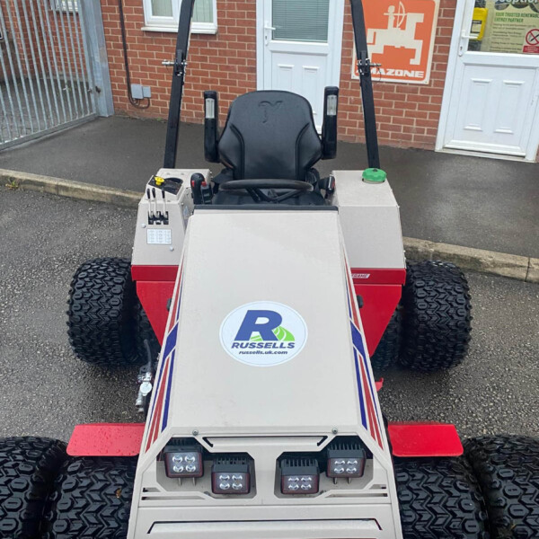 Front view of a small off-road vehicle with a black roll cage, large knobby tires, and a Russell's logo on the hood, parked outside a brick building.