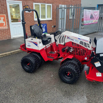 Red and white compact tracked industrial vehicle with a seating area and roll bar, parked outside a brick building near a reception area.
