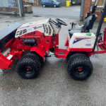 Red industrial machine with large off-road tires, steering wheel, and control panel, parked in a paved lot with a fence and buildings in the background.