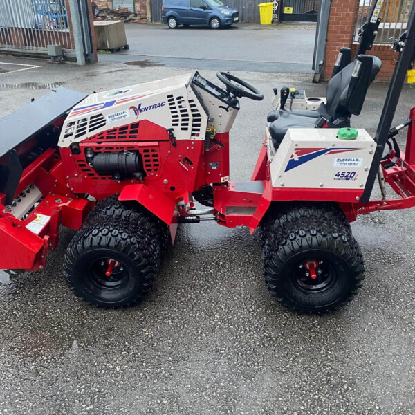 Red industrial machine with large off-road tires, steering wheel, and control panel, parked in a paved lot with a fence and buildings in the background.