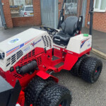 Red-and-white industrial ride-on tractor with large off-road tires and a protective roll bar, parked outside a gated brick building; label stickers visible on the side.