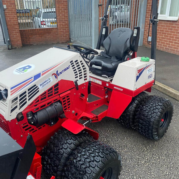 Red-and-white industrial ride-on tractor with large off-road tires and a protective roll bar, parked outside a gated brick building; label stickers visible on the side.