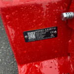 Close-up of a red metal surface with water droplets, a black plate showing a QR code and the serial number HQ682-AA08712, and a chrome bolt nearby.