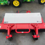 Red agricultural mower attachment with a silver top cover labeled 'Tough Cut HQ682' on a red frame, parked on wet asphalt with green machinery in the background.