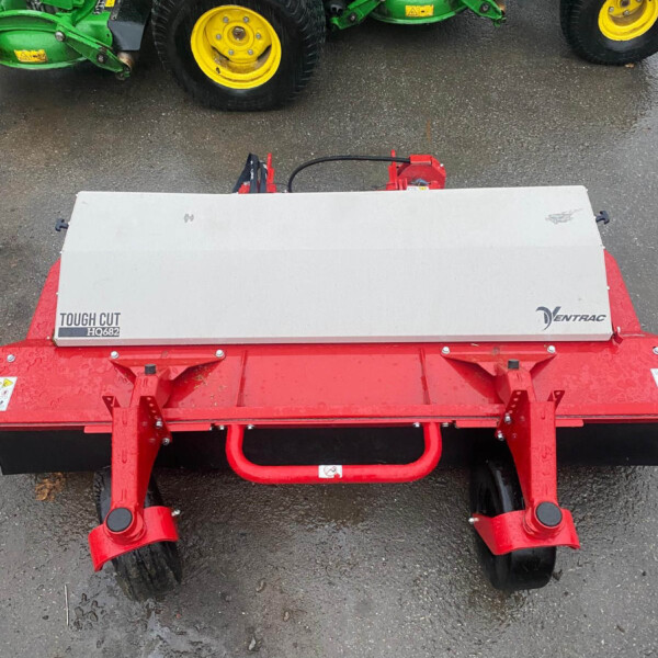 Red agricultural mower attachment with a silver top cover labeled 'Tough Cut HQ682' on a red frame, parked on wet asphalt with green machinery in the background.