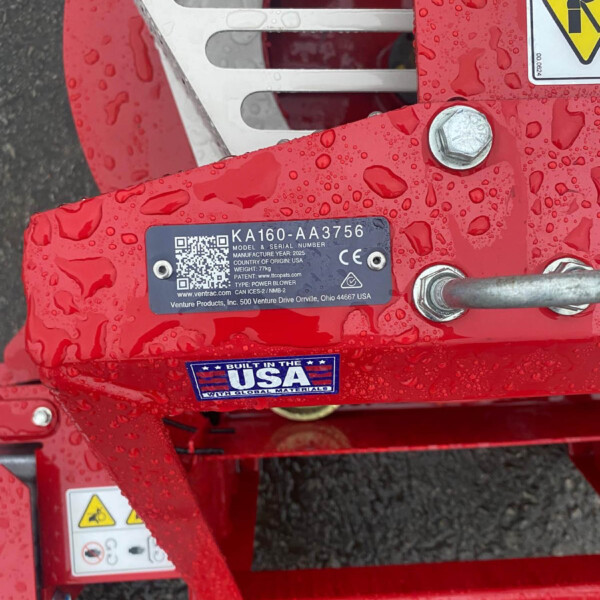 Close-up of a red industrial machine frame with a metal grate, warning stickers, and a blue model label KA160-AA3756 with a QR code, covered in water droplets.
