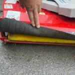 A hand pressing down on a red, wet agricultural roller with warning labels and a yellow bar beneath it in an outdoor setup