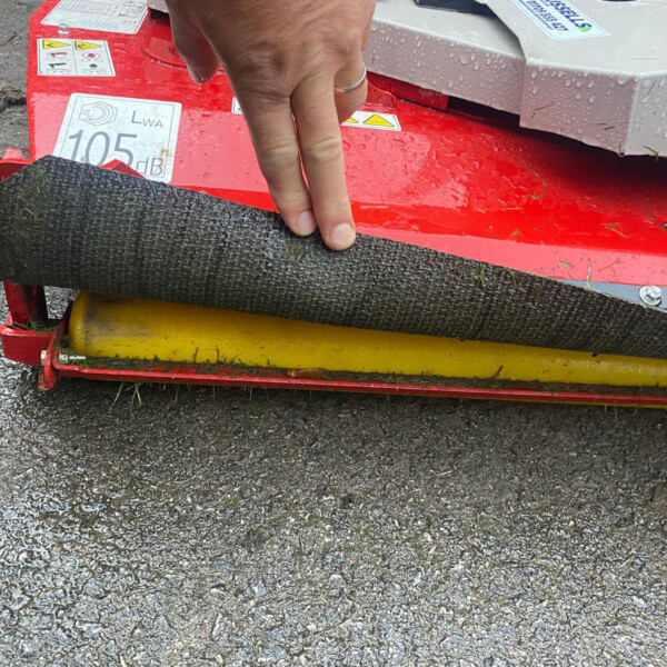A hand pressing down on a red, wet agricultural roller with warning labels and a yellow bar beneath it in an outdoor setup