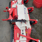 Red agricultural mower attachment with a gray cover, viewed from above on wet pavement, showing warning labels and hydraulic hoses.