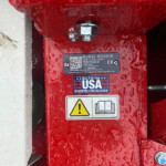 Red lawn mower deck close-up showing a bolt and safety label with a yellow warning icon and a manual icon; surface is wet with droplets.