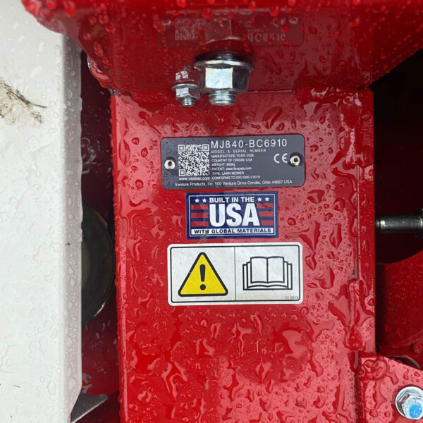 Red lawn mower deck close-up showing a bolt and safety label with a yellow warning icon and a manual icon; surface is wet with droplets.