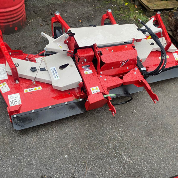 Red agricultural mower attachment with hydraulic hoses on an asphalt surface in a storage yard, showing warning labels and metal guards.