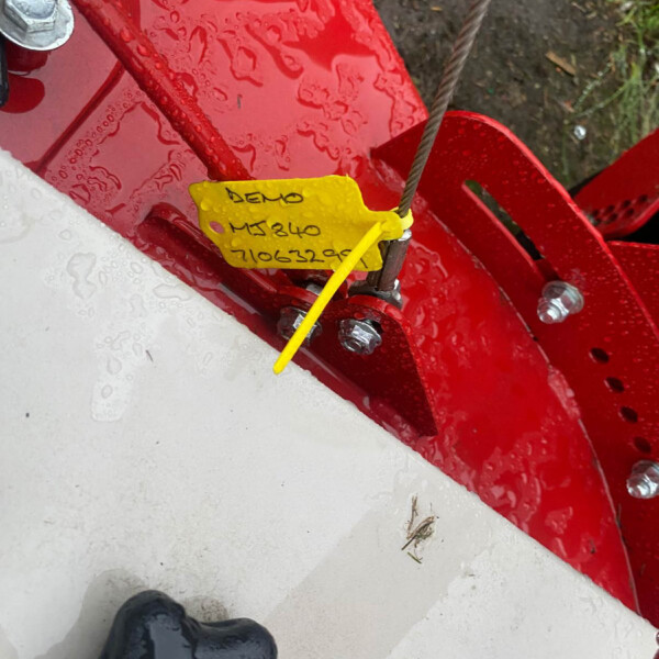Close-up of a red metal frame with a yellow handwritten tag attached by a wire, a steel cable running nearby, water droplets on the metal, and part of a black boot in the lower left.