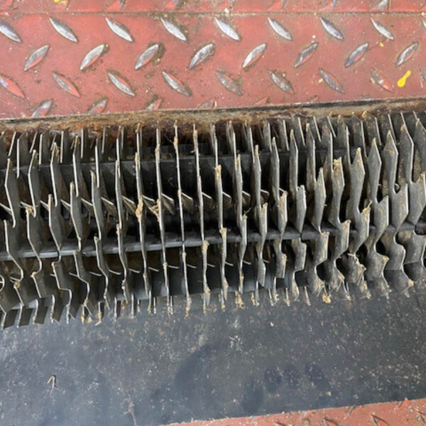 Close-up of a red, diamond-plate metal surface above a row of dark, jagged metal fins or teeth, with some rust and debris.