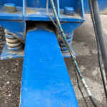 Blue industrial machinery close-up showing springs, hoses, and dirt on gravel ground as part of agricultural equipment.